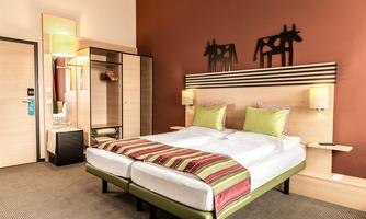 A modern hotel room with a large bed and colorful cushions. The walls are painted in warm colors, and there is a wardrobe as well as a stylish lighting accent.