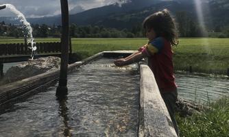 A child plays by a water basin in a picturesque landscape. In the background, green meadows and mountains can be seen under a cloudy sky.