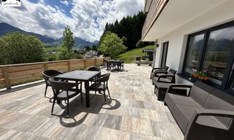 A spacious terrace with modern furniture and a view of a green landscape. The sky is partly cloudy and it's inviting.
