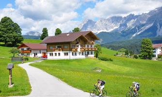 A beautiful country house in a green landscape with mountains in the background. Two cyclists are riding on a quiet road.