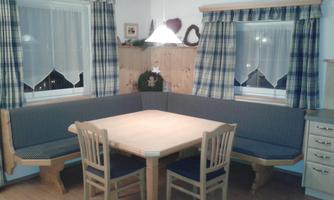 A cozy dining corner with a large tabletop and blue seat cushions. The windows are decorated with checked curtains.