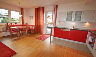 A modern kitchen with red accents and a dining area. Large windows provide plenty of natural light.
