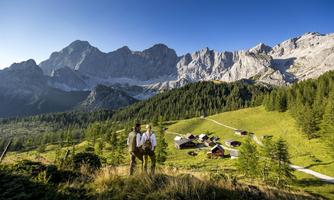 Two men in traditional attire stand in a picturesque mountainous landscape. In the background, there are high mountains and a small Alpine settlement visible.
