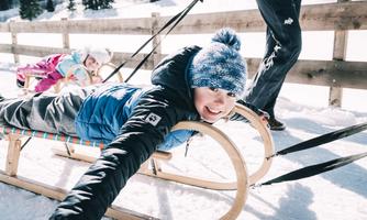 A child is happily lying on a sled in the snow and smiling. In the background, there is a wooden railing and another person visible.