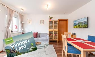 A modern living space with a cozy sofa and a dining table. In the foreground lies a summer brochure that provides information about recreational activities.