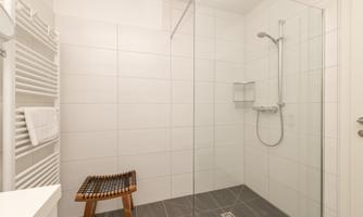 A modern bathroom with a glass shower and white tiles. There is a wooden chair and towels are neatly placed.