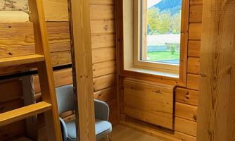 A cozy room with wooden paneling and a window that offers a view of the mountains. In the room, there is a bed, a ladder, and a chair.