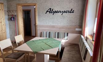 A cozy dining area with wooden decor and a green table. On the wall is the inscription "Alpenperle."