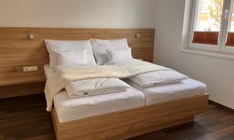A modern bedroom with a double bed and wooden decor. The walls are bright and the room has large windows that let in plenty of daylight.