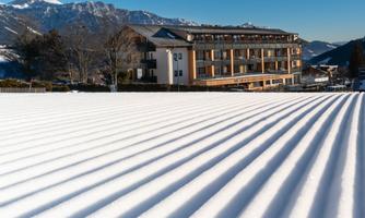 A snow-covered field with even lines in the foreground. In the background, a hotel can be seen against a mountain backdrop and a clear blue sky.