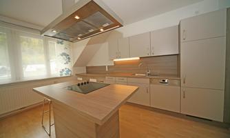 A modern kitchen with light wood surfaces and a cooking island. Large windows ensure plenty of daylight.