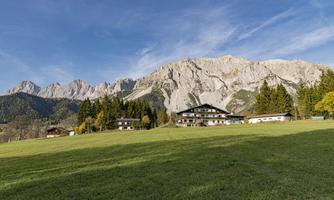 A picturesque landscape with green meadows and majestic mountains in the background. In the foreground, there are some traditional houses surrounded by trees.