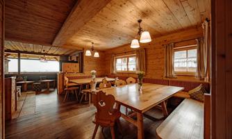 A cozy wooden room with a large table and benches. The warm lights and windows create an inviting atmosphere.