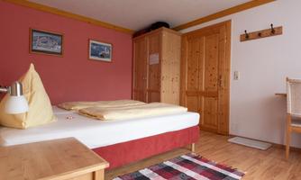 A cozy bedroom with a double bed and wooden furniture. Pictures hang on the wall, and the floor is covered with a colorful rug.
