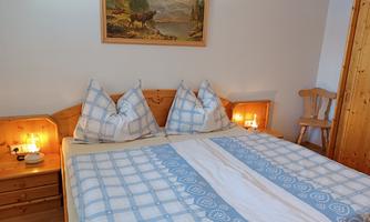 A cozy bedroom with a double bed and blue pillows. Evening light shines from the nightstands and a wooden chair.