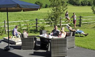A cozy terrace with a table and chairs, surrounded by green meadows. People spend time outdoors, enjoying the sun and relaxing.