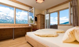 A cozy bedroom with a large window offering a view of the mountains. The furnishings are made of light wood, and the room feels inviting and bright.