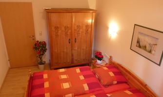 A cozy bedroom with a double bed and colorful bedspreads. A wooden wardrobe and decorative plants complete the room.