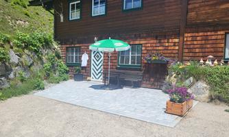 A cozy wooden house with a small seating area and a green sun umbrella. Blossoming plants and a well-maintained terrace enhance the serene atmosphere.
