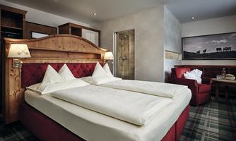 A cozy hotel room with a large double bed and red furniture. There is a quiet area with an armchair and a modern bathroom.