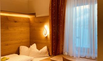 A cozy bedroom with wooden paneling and a single bed. There is a desk with a chair and pleasant light by the window.