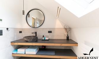 A modern bathroom with a wooden shelf and a round mirror. On the shelf are a washbasin and some towels in various colors.