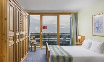 A cozy hotel room with a large bed and a wooden wardrobe. The view from the window offers a beautiful view of the mountains.