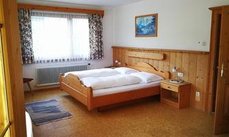 A bright bedroom with a double bed and wooden paneling. The room has large windows with curtains and a cozy atmosphere.