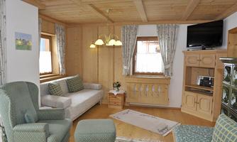 A cozy living area with wood paneling and bright lighting. The room features a sofa, armchair, and a television, perfect for relaxing.