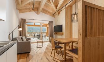 A modern, cozy living room with wooden furniture and large windows. The room offers a view of the terrace and the surrounding nature.