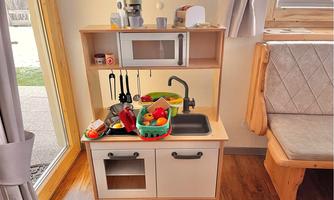A small play kitchen with various toys, a sink, dishes, and a cooktop. The kitchen is located next to a window with a view of the outdoors.