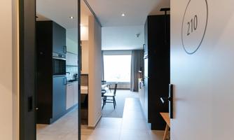 A modern hotel room entrance overlooking a bright kitchen and living area. The walls are in neutral colors, and the light is friendly and inviting.