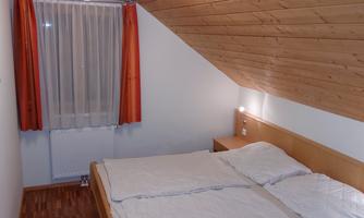 A cozy bedroom with a double bed and wooden paneling. The windows are decorated with light curtains and orange-colored drapes.