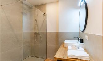 A modern bathroom with a glass shower and a wooden washbasin countertop. The walls are in neutral tones and there are fresh towels on the shelf.