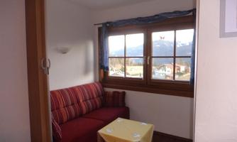 A cozy room with a red sofa and a small table. Large windows offer a view of the mountains and the surroundings.