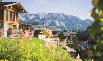 A picturesque residential area with wooden chalets and magnificent mountains in the background. Colorful flowers adorn the front area, giving the scene a charming appearance.