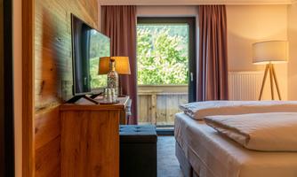 A modern hotel room with two beds and a television. The view looks out onto a balcony with green trees in the background.