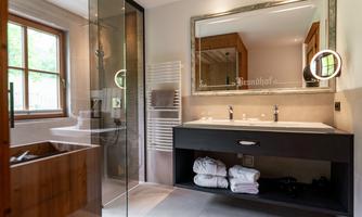 A modern bathroom with a large shower and a double sink. On the right, towels are neatly stacked, and a large mirror reflects the light from the windows.