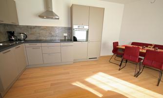 A modern kitchen with bright cabinets and a dining table with red chairs. The room is well-lit and has a warm wooden floor.