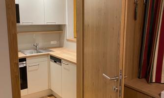 A modern kitchen with white cabinets and wooden flooring. The door opens into an inviting space.