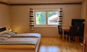 A bright room with two beds, a table, and a television. Large windows with curtains provide plenty of daylight.
