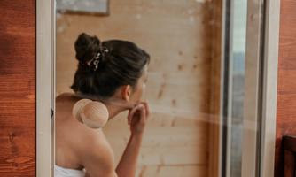 A woman is sitting in a sauna and looking thoughtfully outside. She is wearing a towel and the walls are made of wood.