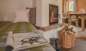A cozy corner with a padded bed and a fireplace. On the table lie a newspaper and glasses, while next to it stands a basket of wood and some boots.