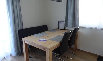 An elegant dining area with a wooden table and several chairs. The windows are decorated with light curtains, and a modern pendant light hangs.