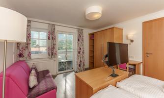 A modern room with a pink sofa, a desk, and a television. Large windows with curtains allow plenty of light in.