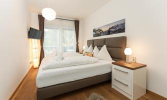 A modern bedroom with a large double bed and bright walls. Next to the bed are nightstands, and a large window front provides plenty of daylight.
