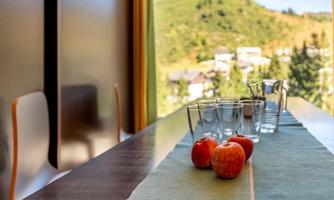 An elegant table with apples and glasses, offering a beautiful view of the mountains. The atmosphere is bright and inviting.