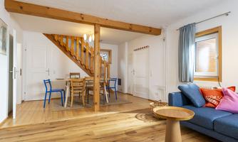A bright living room with wooden floors and a stylish staircase. A blue sofa, a table, and chairs create a cozy atmosphere.