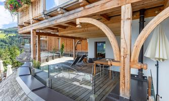 A modern terrace with wooden cladding and glass railing overlooks a picturesque mountain landscape. The architecture combines traditional and contemporary elements.