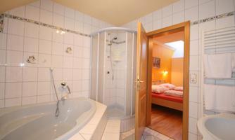 A modern bathroom with a round bathtub and a shower cabin. In the background, a bedroom with two beds is visible.